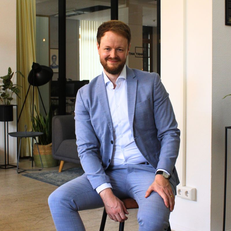Marketingberater im hellblauen Anzug sitzt auf Hocker an Konferenzraum lächelt in die Kamera