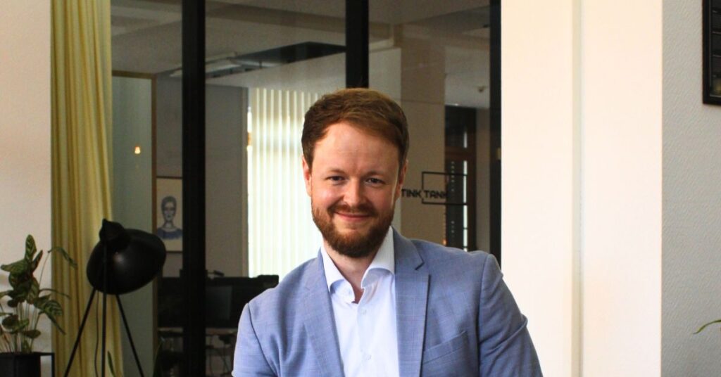 Marketingberater im hellblauen Anzug sitzt auf Hocker an Konferenzraum lächelt in die Kamera